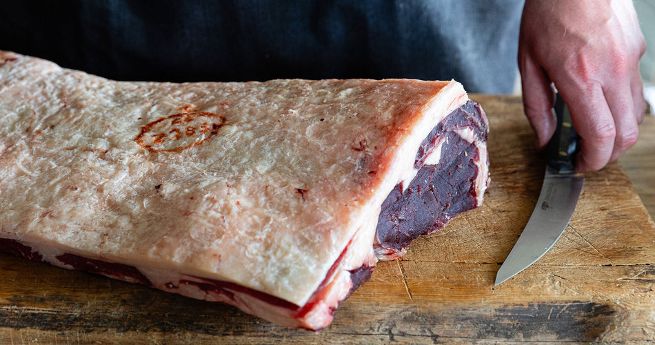 Raw whole grass-fed beef sirloin rack, bone-in, with fat cap, on a butcher’s block.