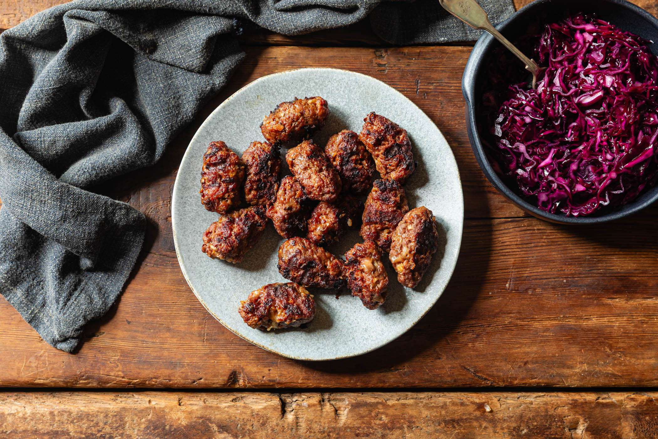 A plate of browned, crispy-fried sausage links arranged on a light gray ceramic plate, set on a rustic wooden table next to a bowl of shredded red cabbage and a dark gray cloth napkin.