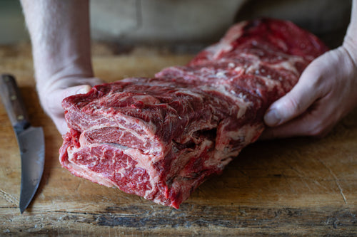 Raw beef cut chuck eye roll on a wooden cutting board with hands holding it, knife on the side