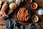 A rustic overhead flat-lay of seasoned ground meat in a black bowl, surrounded by raw ingredients including sliced bacon, a whole onion, garlic, spices in small bowls, and a dark gray cloth on a wooden table.