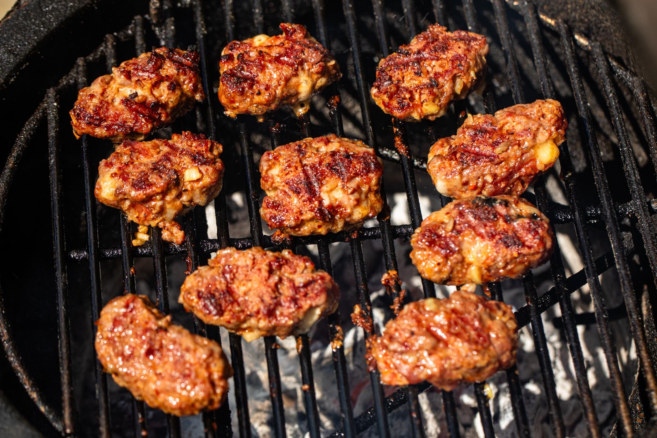 Several browned meatballs or fritters cooking on a black grill grate over white coals.