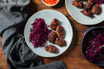 Overhead food styling on a wooden table with a light gray plate holding several browned meatballs next to shredded purple cabbage, with additional plates and bowls of food partially visible.