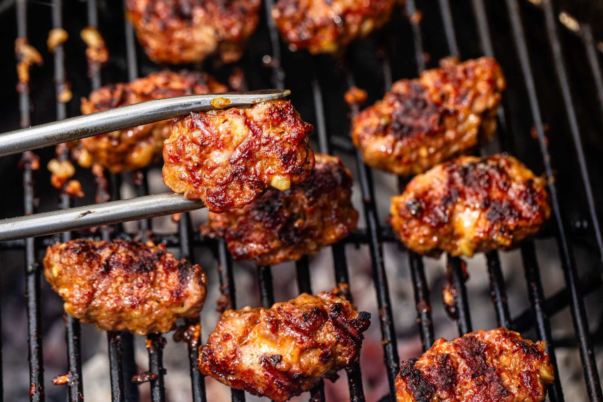 Grilled meatballs on a black grill grate with metal tongs holding one piece, shown in a close-up food scene.