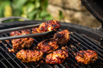 Grilled meatballs on a black barbecue grill with tongs holding one piece, set against a blurred outdoor background.