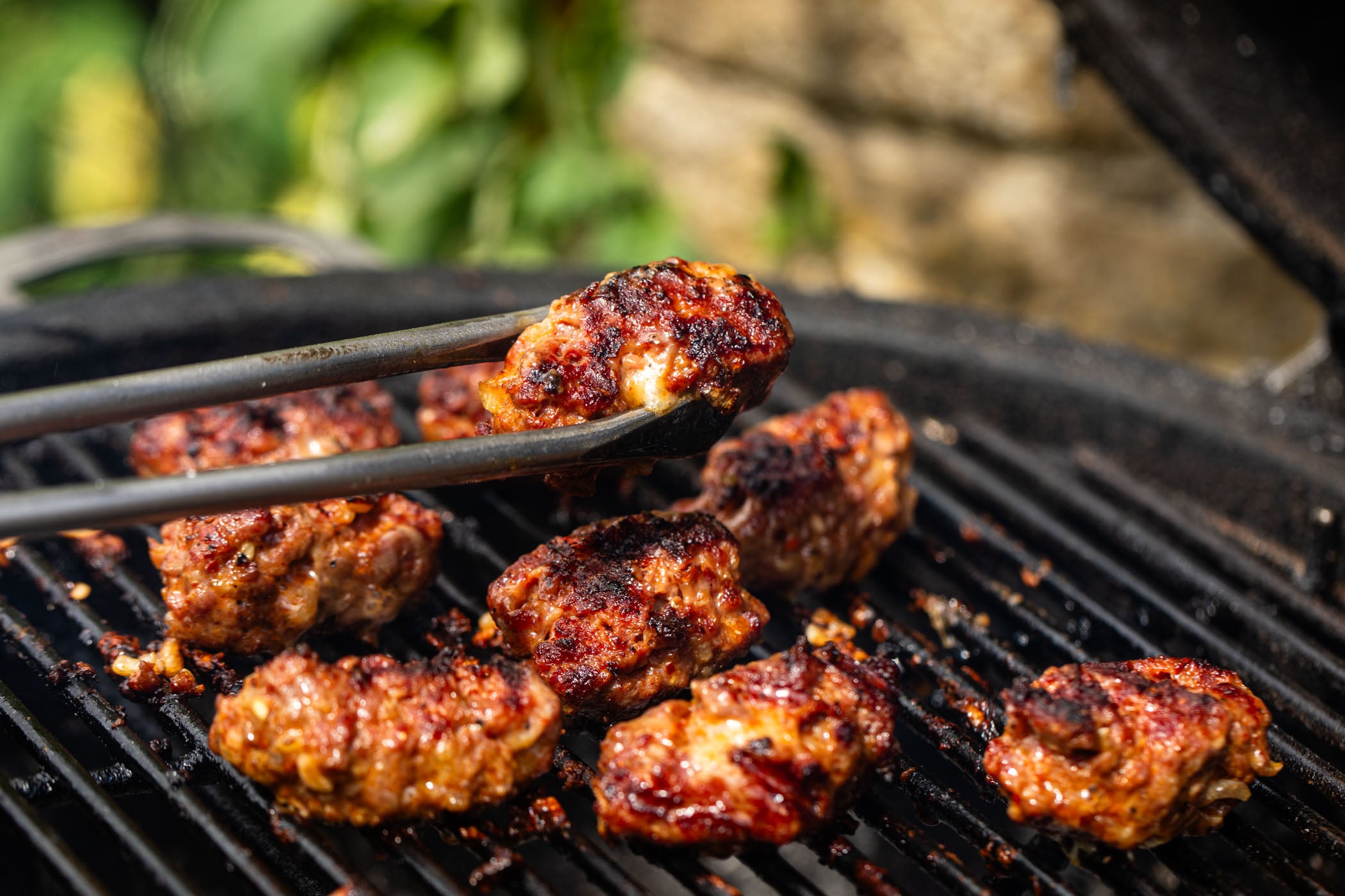 Grilled meatballs on a black barbecue grill with tongs holding one piece, set against a blurred outdoor background.