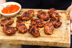 Grilled meatballs on a wooden serving board with a small bowl of orange-red dipping sauce, held by hands in a real setting.
