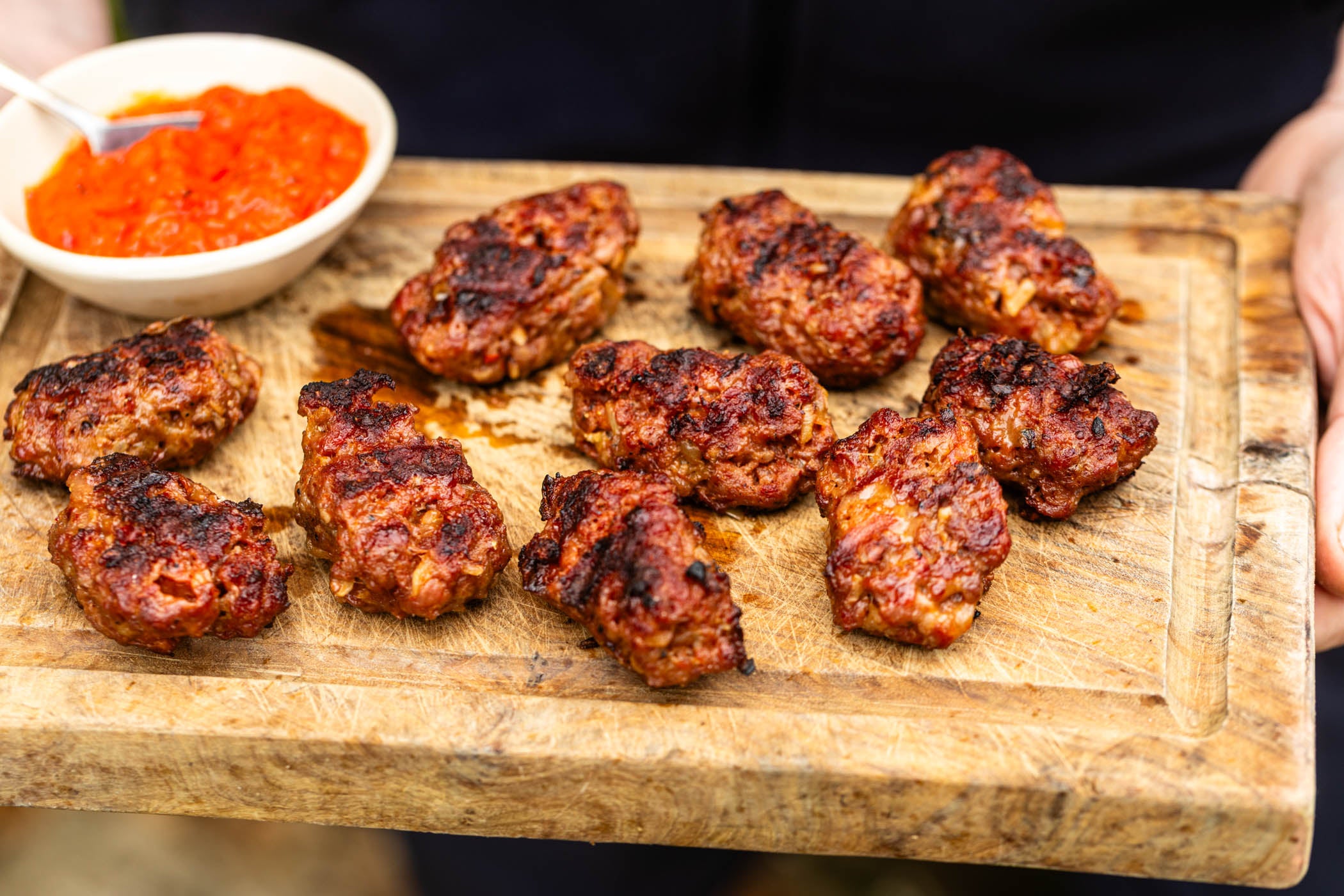 Grilled meatballs on a wooden serving board with a small bowl of orange-red dipping sauce, held by hands in a real setting.