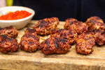 Grilled meatballs with charred spots served on a wooden cutting board, with a small bowl of red sauce in the background.
