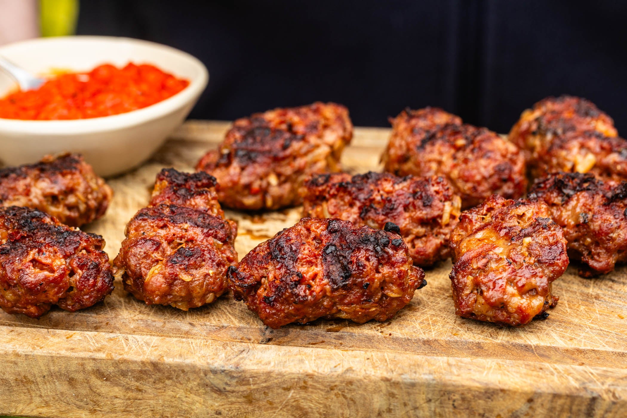 Grilled meatballs with charred spots served on a wooden cutting board, with a small bowl of red sauce in the background.