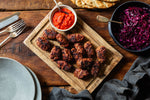 Grilled meatballs served on a wooden board with a bowl of red sauce, a bowl of shredded red cabbage, and flatbread on a rustic wooden table.