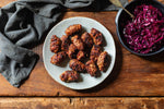 A plate of browned, crispy-fried sausage links arranged on a light gray ceramic plate, set on a rustic wooden table next to a bowl of shredded red cabbage and a dark gray cloth napkin.