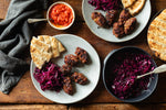 Overhead flat-lay of a prepared meal on a rustic wooden table, featuring grilled meatballs on plates with pita bread, red cabbage salad in a bowl, and a small bowl of red sauce.