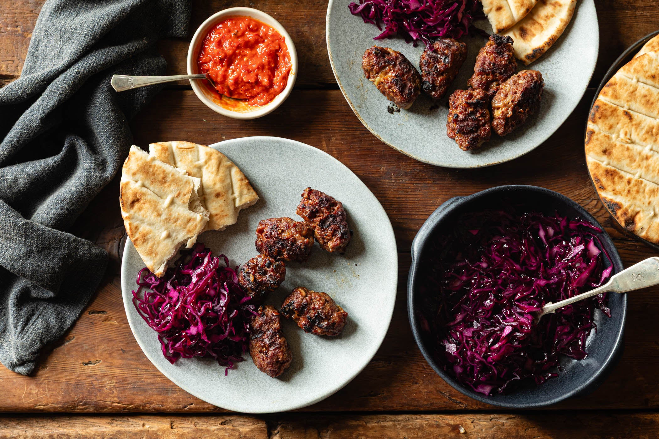 Overhead flat-lay of a prepared meal on a rustic wooden table, featuring grilled meatballs on plates with pita bread, red cabbage salad in a bowl, and a small bowl of red sauce.